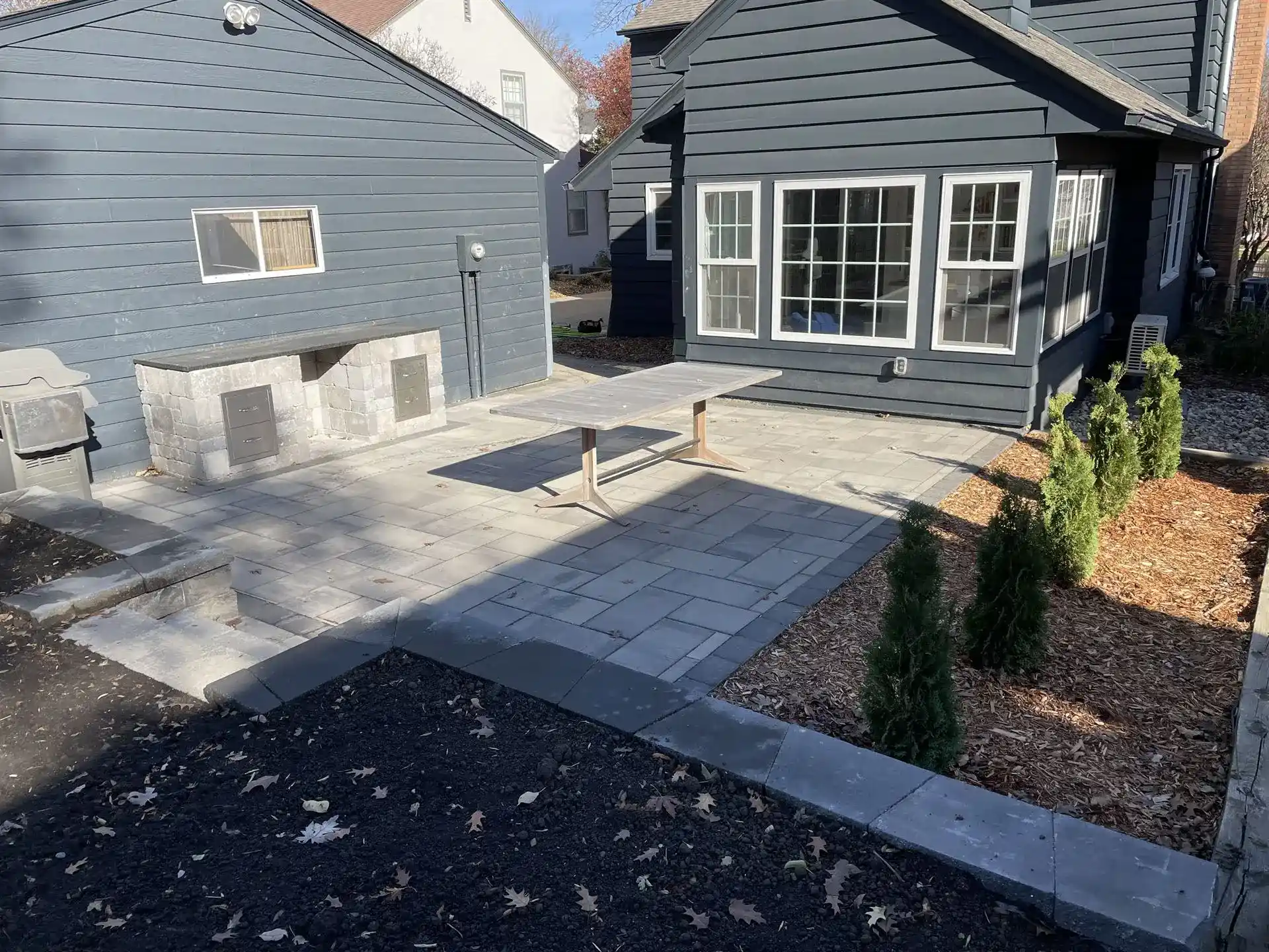Backyard patio beside a dark blue house, featuring a light gray stone paver surface with a simple rectangular table, a built-in stone counter along the garage wall, and a mulched planting bed.