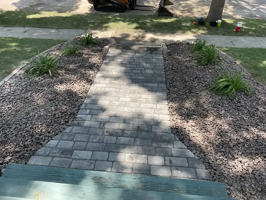 View looking down a newly installed stone walkway made of gray rectangular pavers, bordered on both sides by beds of small brown rocks with several green plants.